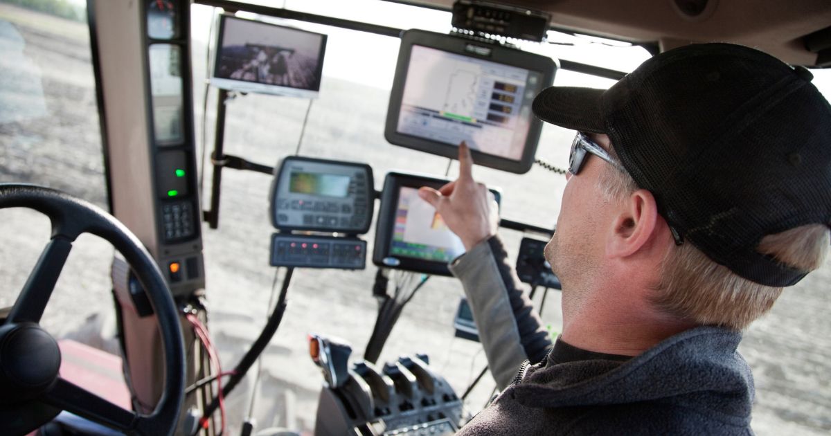 Man in his 30s in a tractor on the farm. It is seeding time in the spring. There are many monitors to his right, this is a hi-tech farming operation.